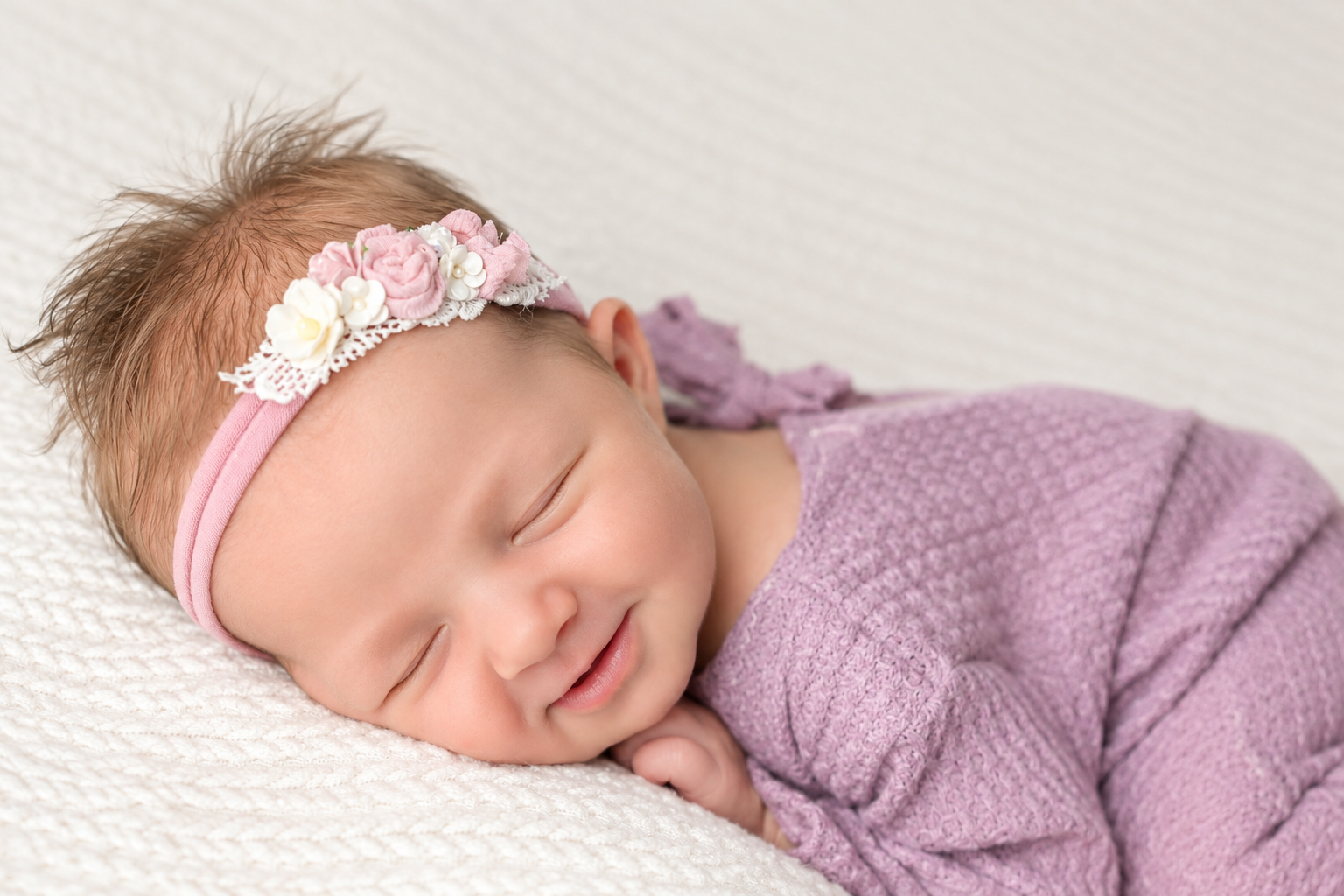 Smiling newborn baby wrapped in purple with a pink floral headband on a white blanket